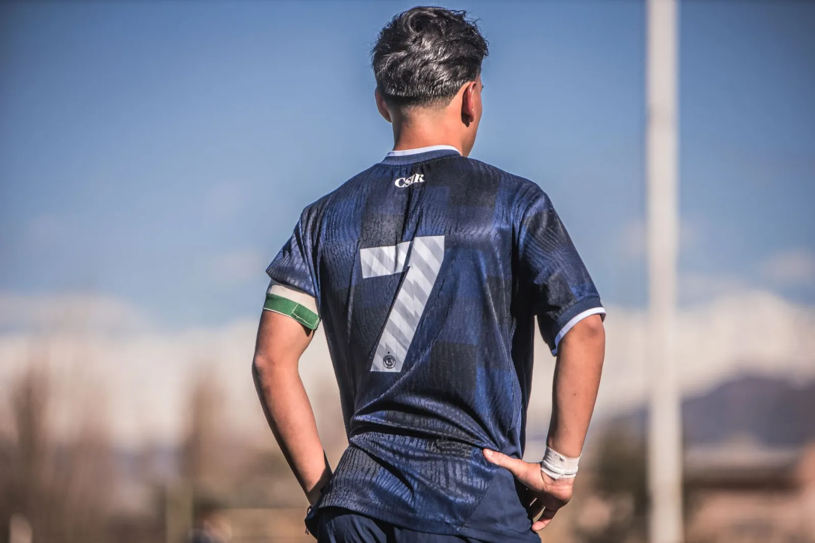 Young soccer player wearing a number 7 blue jersey stands on the field with a clear sky background.