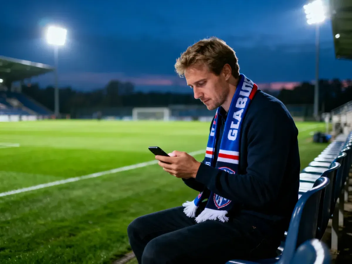 Kibic z szalikiem siedzi na trybunach stadionu i sprawdza wyniki meczu na smartfonie. W tle widać oświetloną murawę boiska.